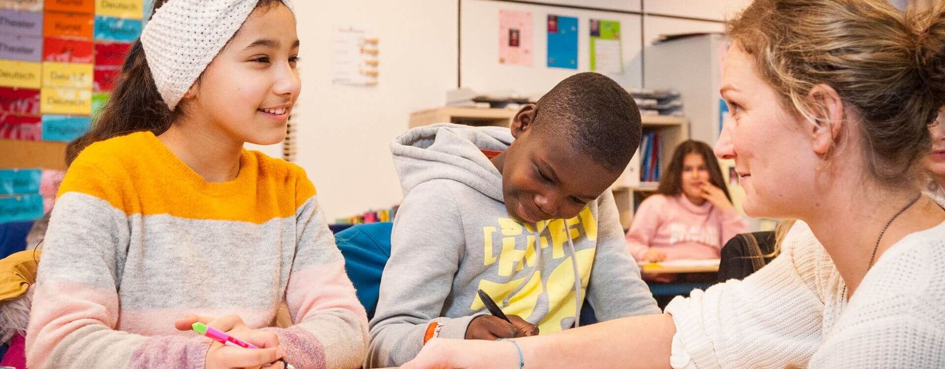 Drei Personen sitzen an einem Tisch in einem Klassenzimmer der Stadtteilschule Mümmelmannsberg, zwei Kinder und eine Erwachsene, die auf ein Heft zeigt.